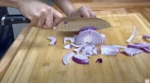 Person slicing red onion on a wooden cutting board with a knife.